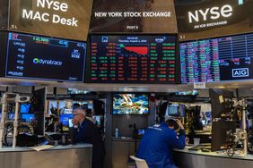Traders work at desks on the floor of the New York Stock Exchange, sitting beneath screens displaying stock data. 