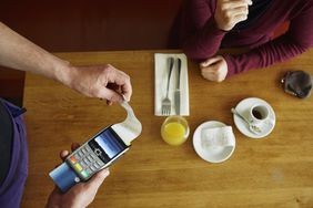 A waiter prints a credit card receipt using a handheld card reader for a customer seated at a cafe table.
