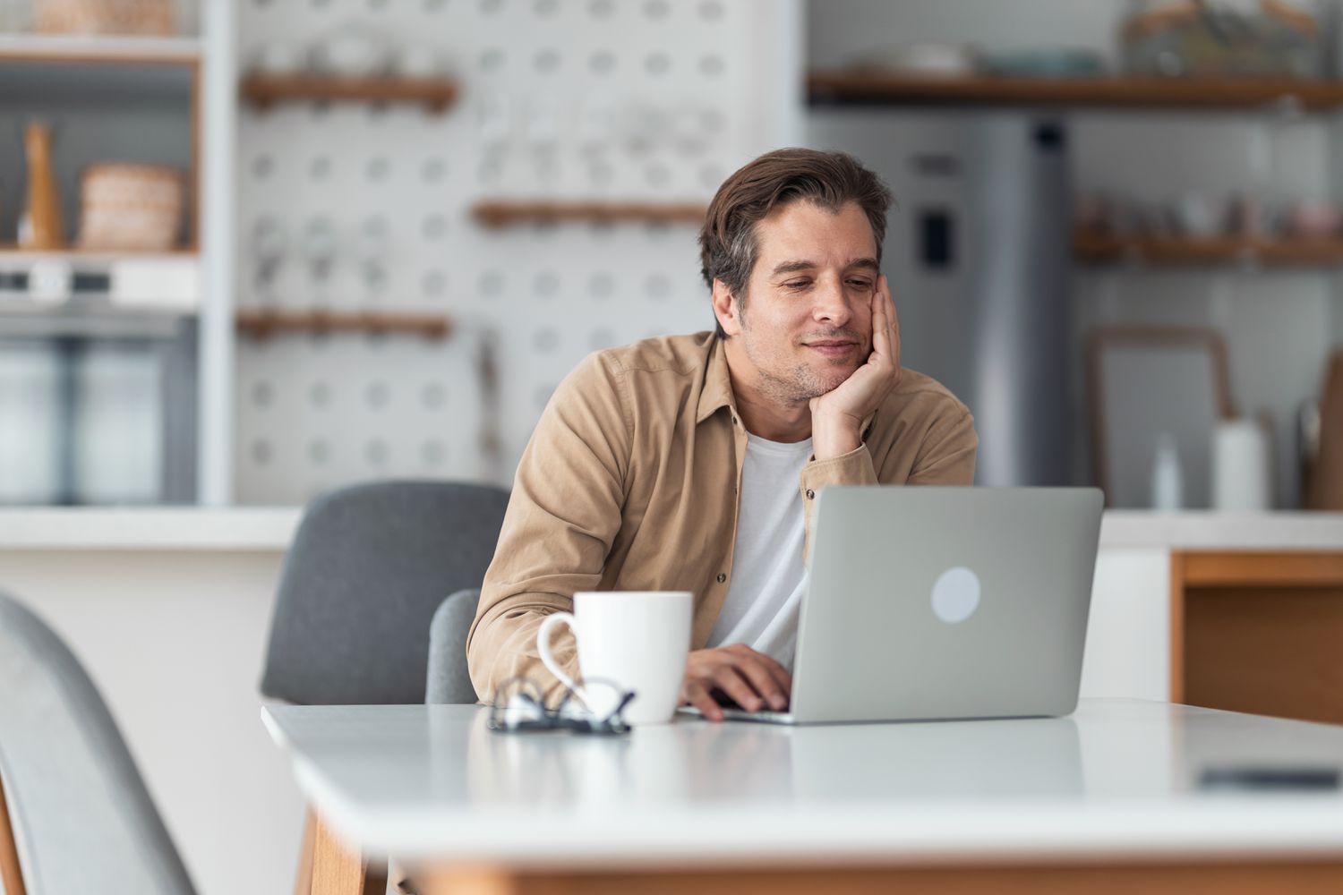 Man in his 30s sitting at his kitchen table and looking happily at something on his laptop