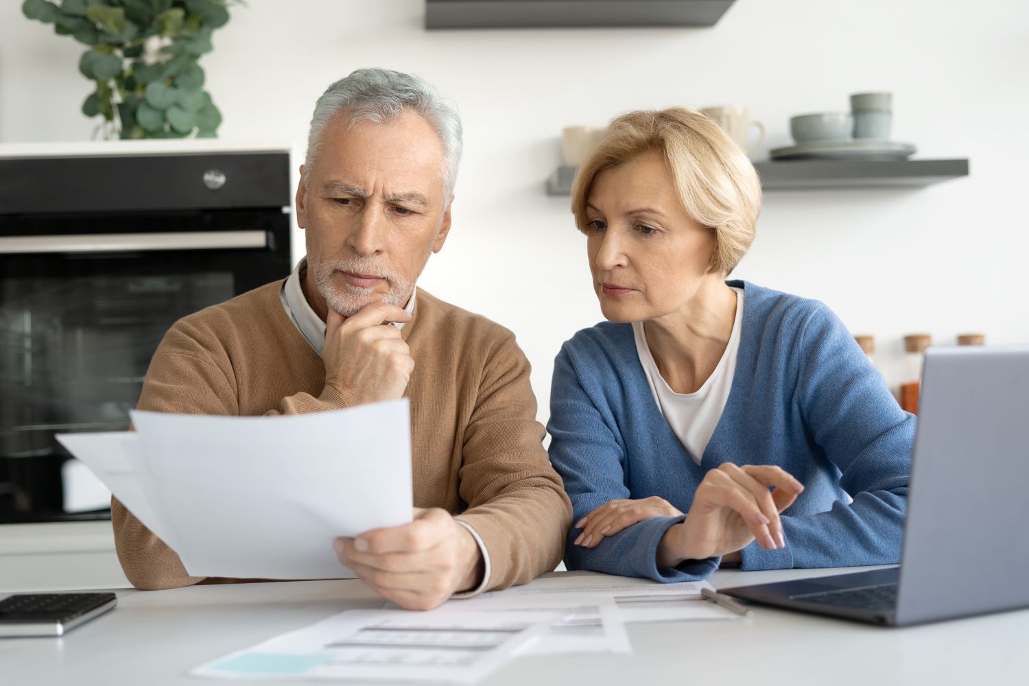 Older couple sitting at their kitchen table and looking together at mortgage documents and a laptop
