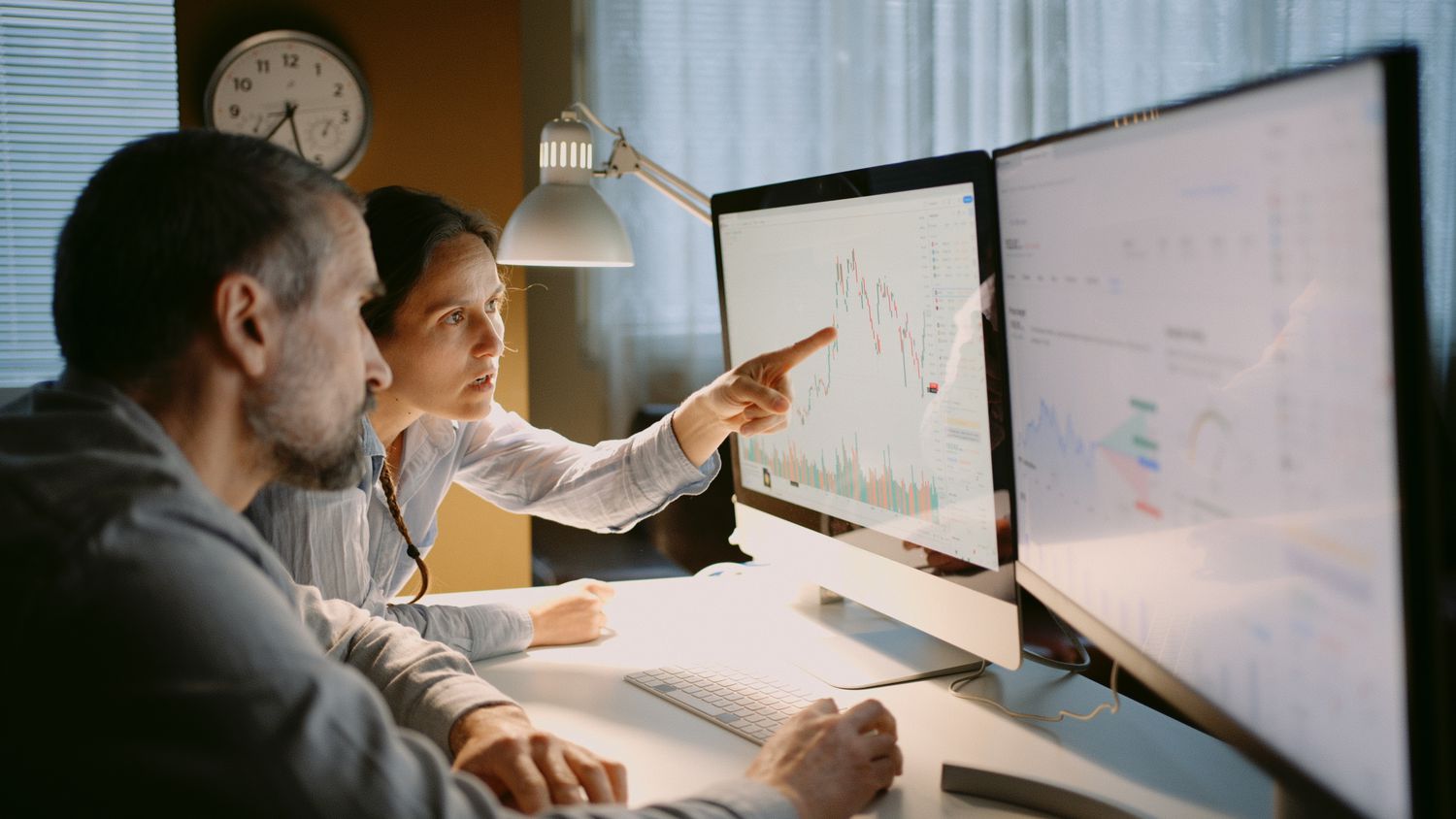 Two traders work at a dest with monitors showing stock data to check trading positions and speculate on market direction.