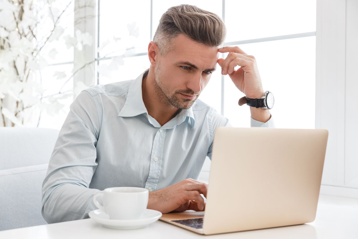 Young man sitting at his kitchen table and seriously pondering something on his laptop