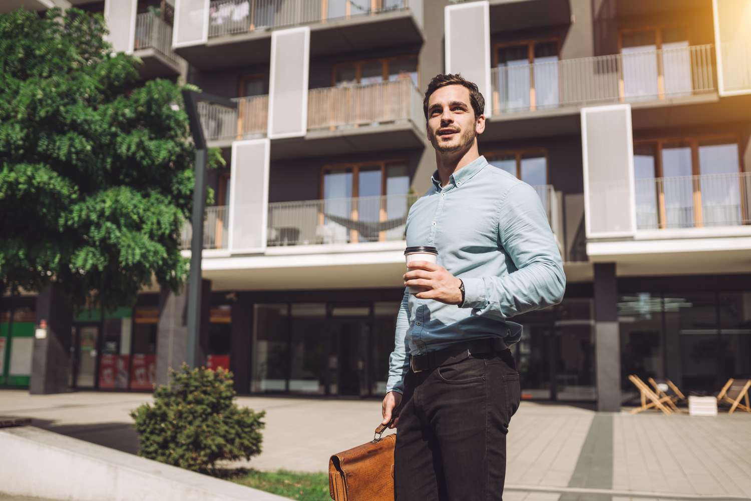 Young man with briefcase and paper coffee cup in front of apartment building