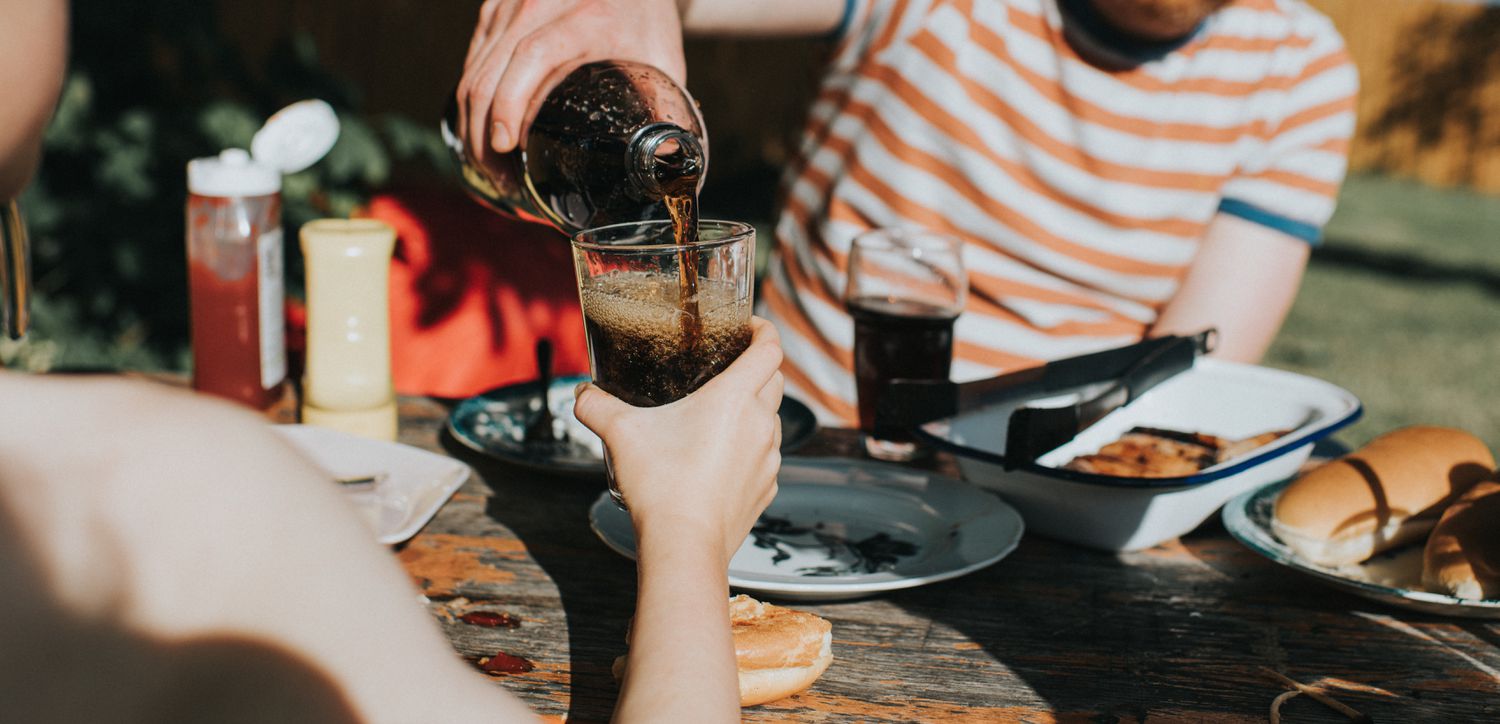A child holds a glass out toward another person, who fills it with a fizzy drink.
