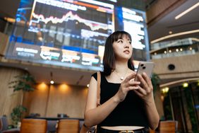 Young woman sanding with her back to a large screen showing market data and charts.