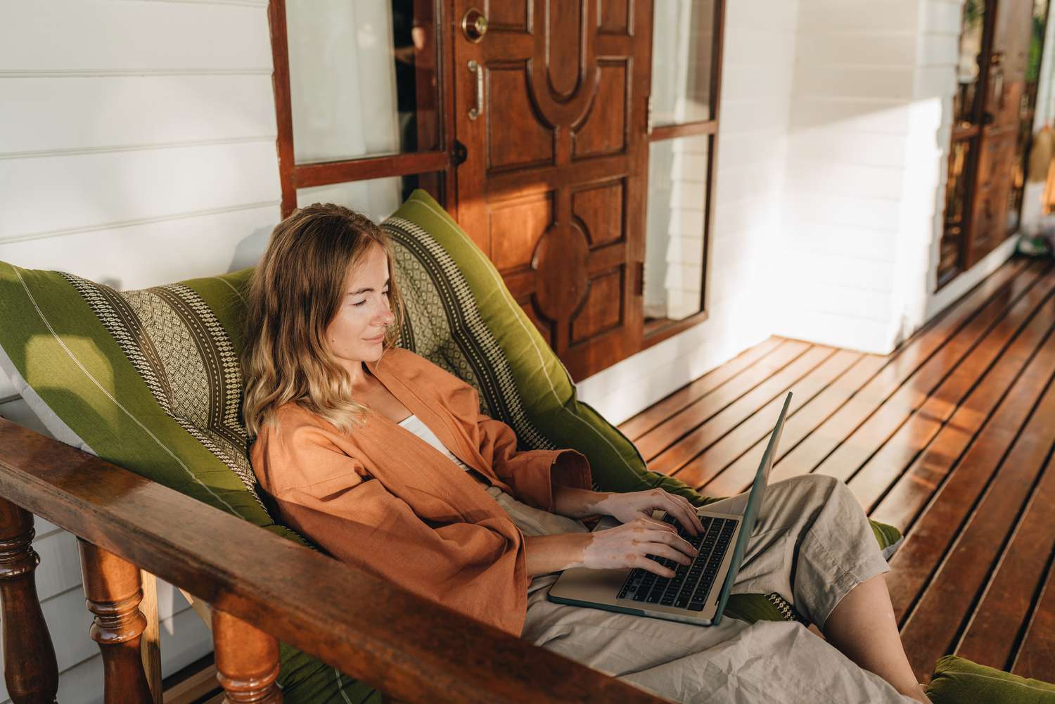 A woman relaxes in a lounge chair on a front porch. She types on the keyboard of the open laptop that rests on her lap. 