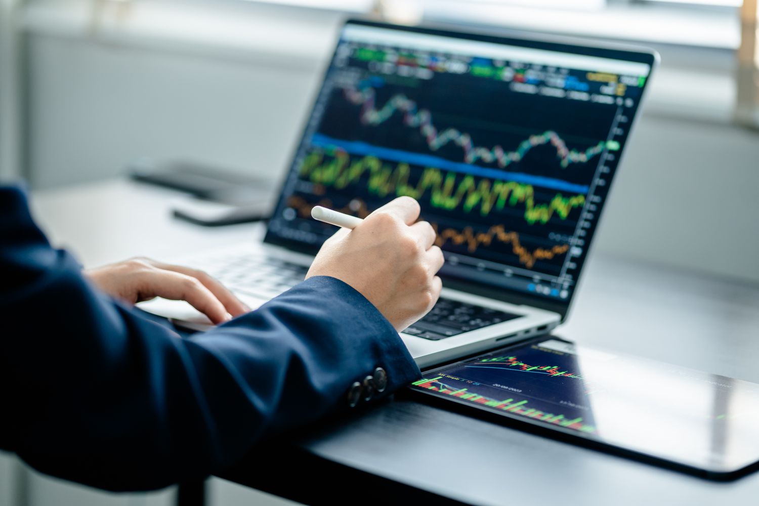A person wearing a suit jacket holds a pen while using a laptop and tablet displaying financial charts.