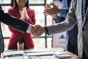Two businessmen reaching acorss a table and shaking hands during a business meeting while coworkers celebrate in background