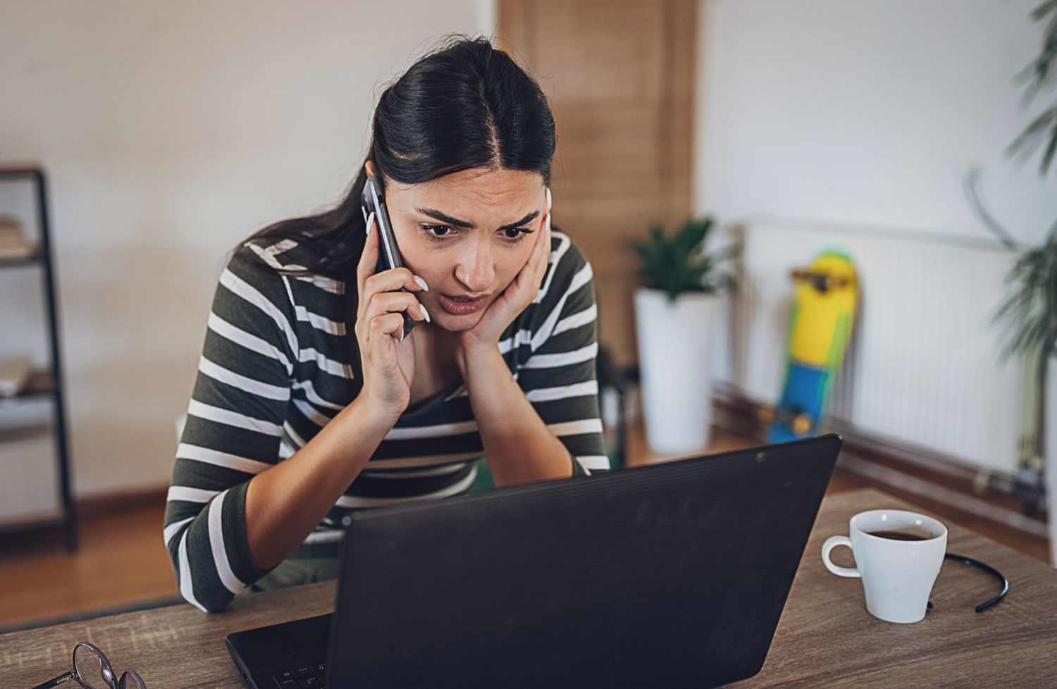 A young person staring anxiously at a laptop, phone in hand.