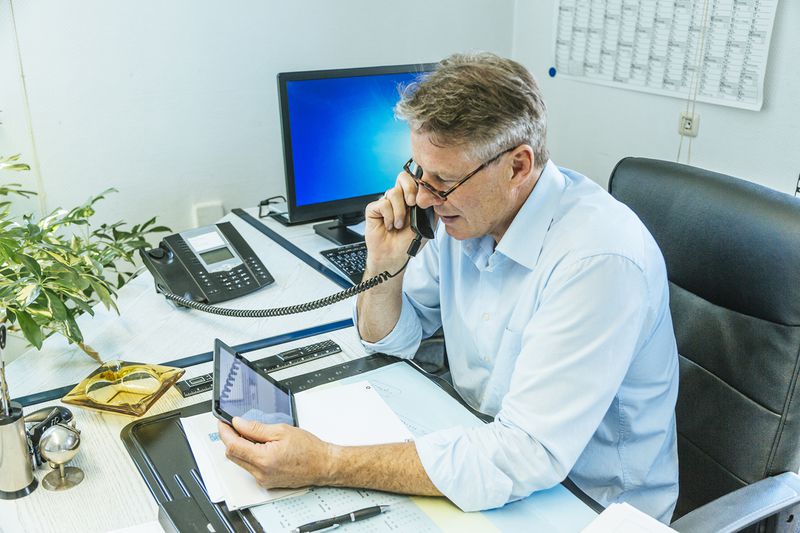Mature Businessman Sitting at His Desk Using a Phone While Holding a Tablett PC