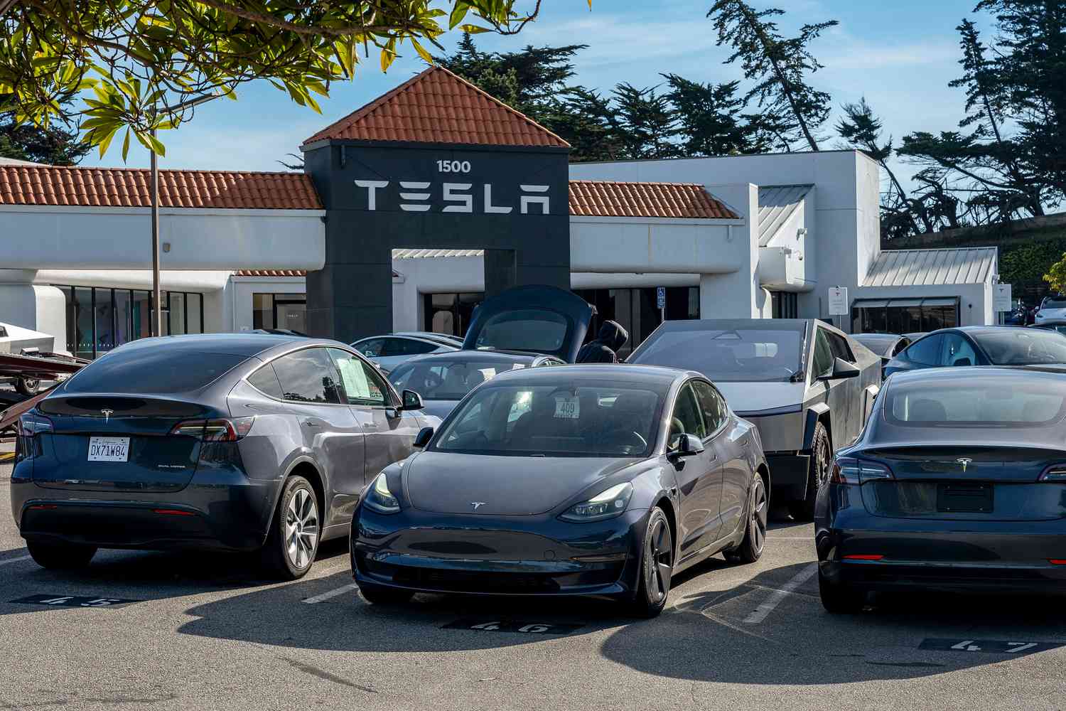 Several Tesla vehicles are seen outside a dealership, with the Tesla logo on the side of a building in the background.