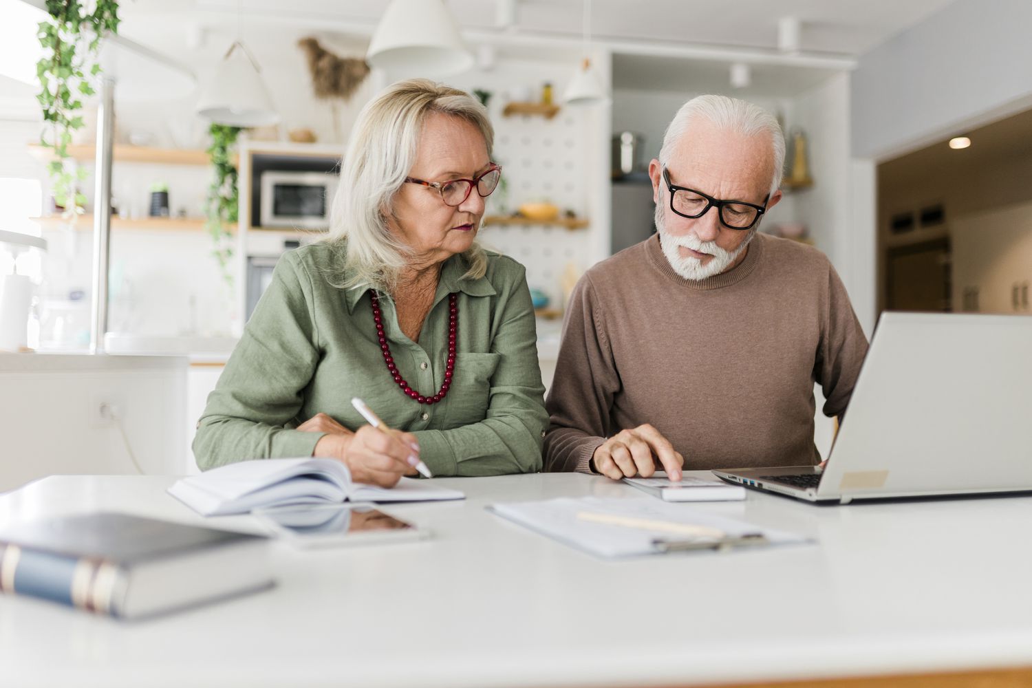 An older adult couple sit at a table using a calculator and laptop as they calculate their possible income from an unfunded pension plan.