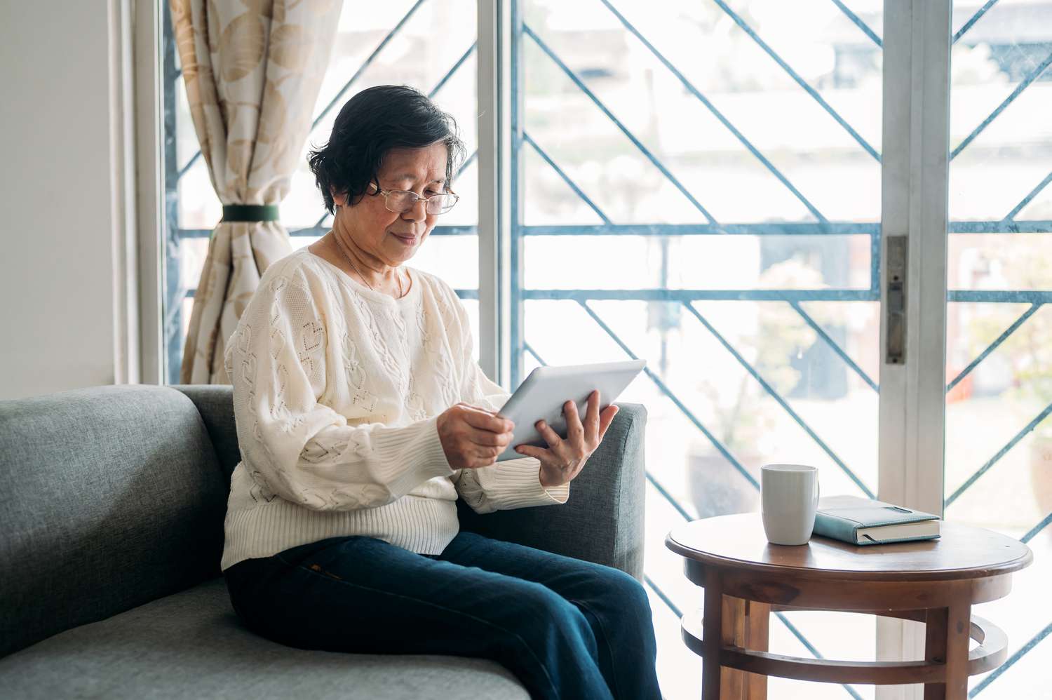 Woman in her 70s at home using a tablet. She is sitting on a grey sofa in front of a window in the living room.