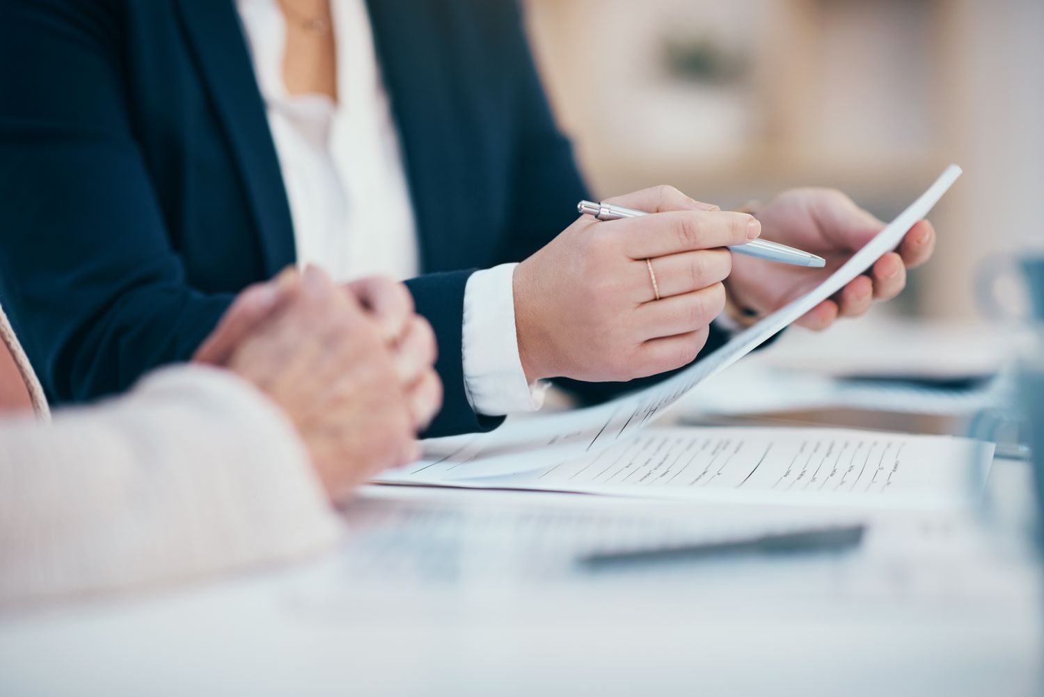 A close up showing the arms and hands of two people at a desk while one of them reads over an insurance policy before signing the document.
