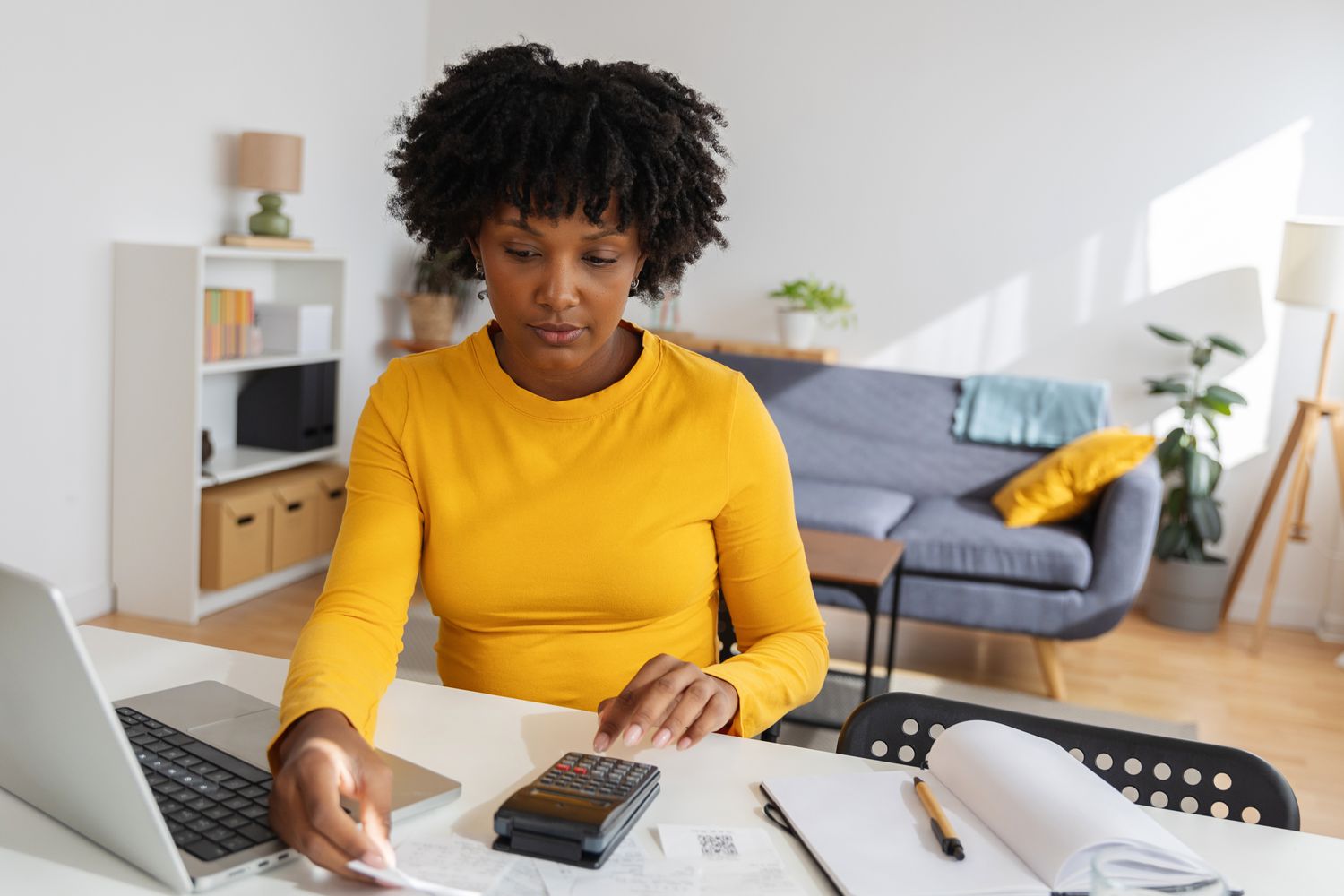 Woman sitting at a desk in a home office using a calculator, with on open laptop next to her.