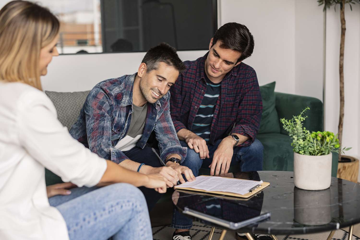 Two men signing paperwork with a woman helping them. They are all sitting in a living room