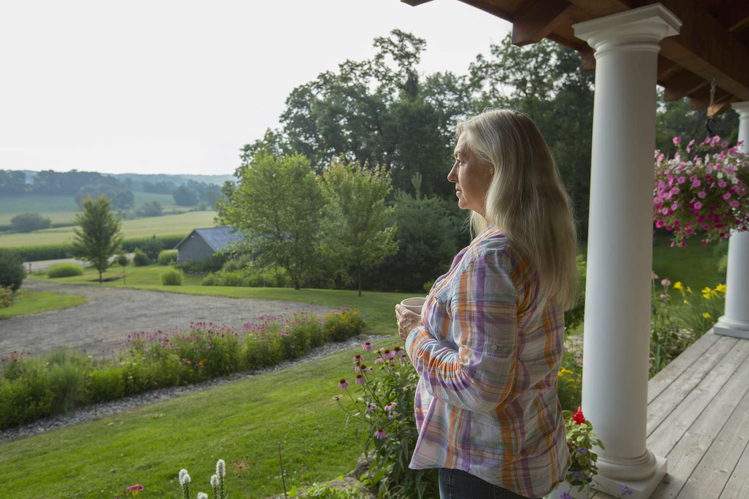 Person with long, light-colored hair holds a mug while standing on the front porch of their home in the Midwest countryside.