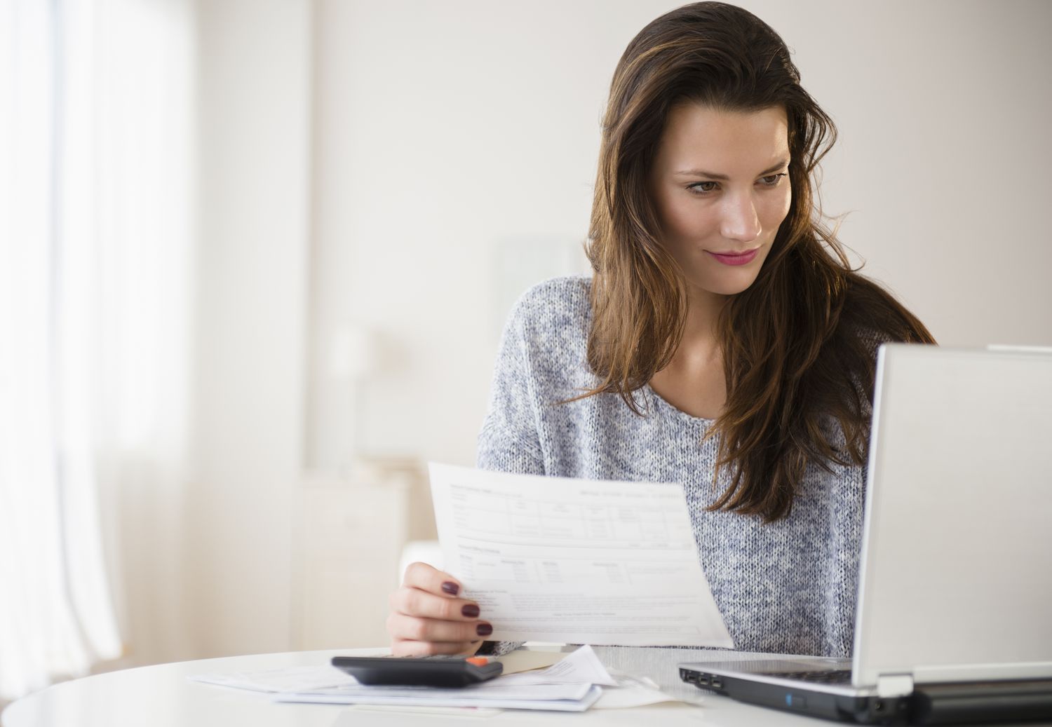 Woman looking at a laptop and holding a piece of paper as a calculator sits on her table.