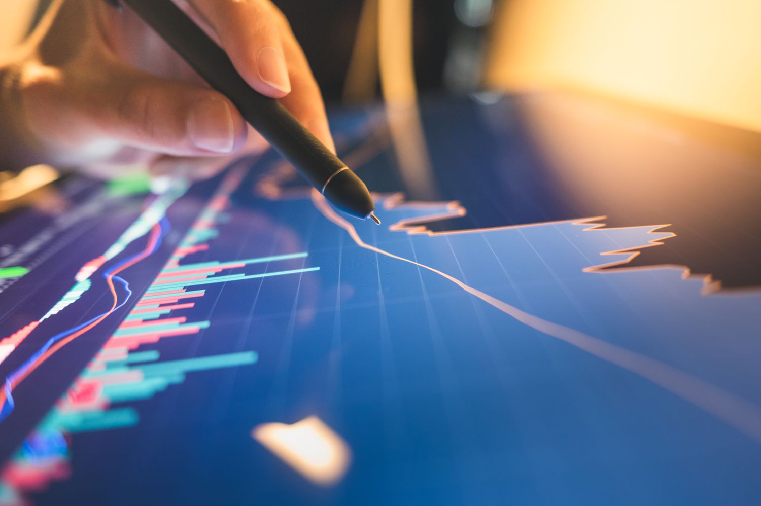 A closeup of a hand holding a pen and pointing to a line on a screen displaying stock data that shows a recovery in price.