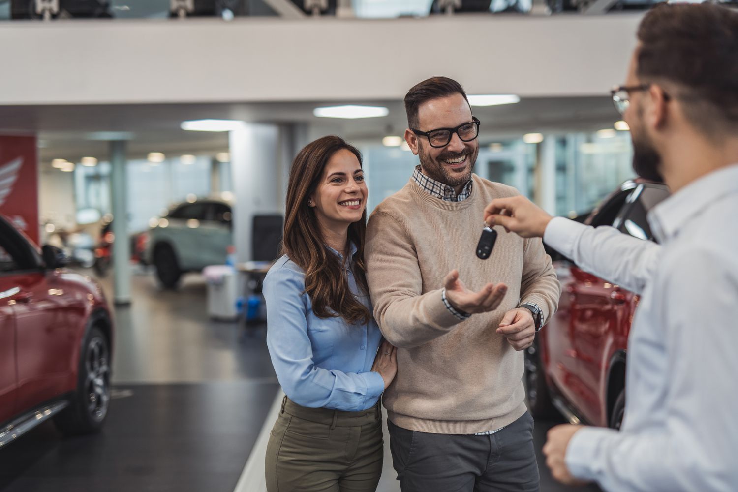 In a car dealership, a happy couple smiles as they receive their new car keys from the salesman.