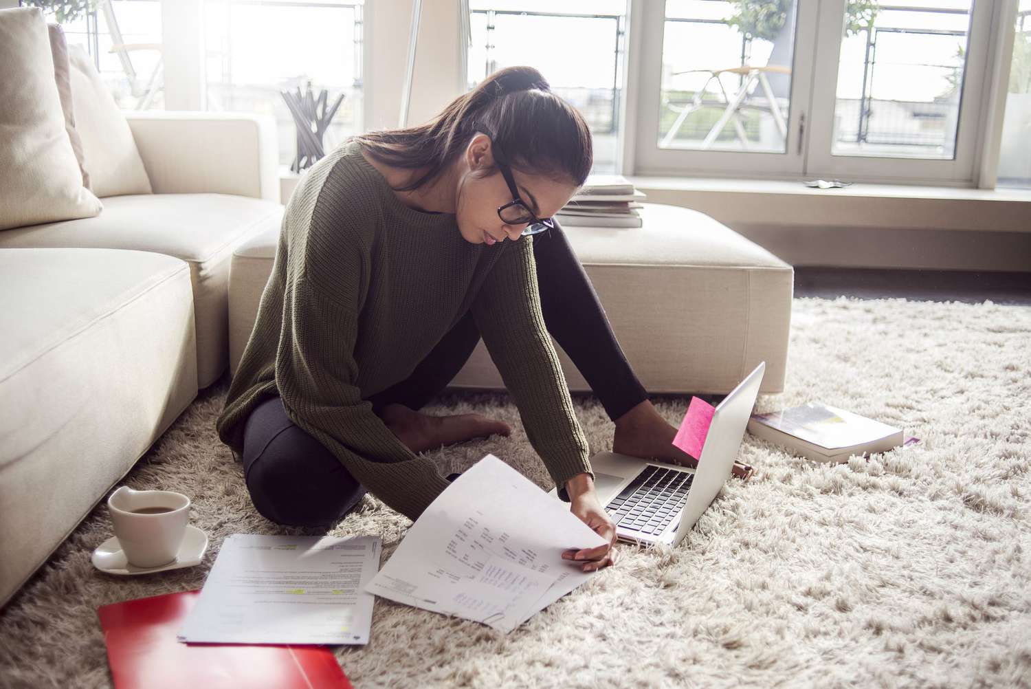 Young woman in glasses reviewing paperwork on the floor of her living room with laptop.