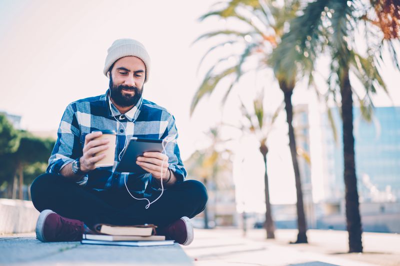 young man in stylish hat watching online webinar in earphones conected to digital tablet.Bearded hipster guy listening audio lesson using touch pad sitting in lotus position with coffee to go