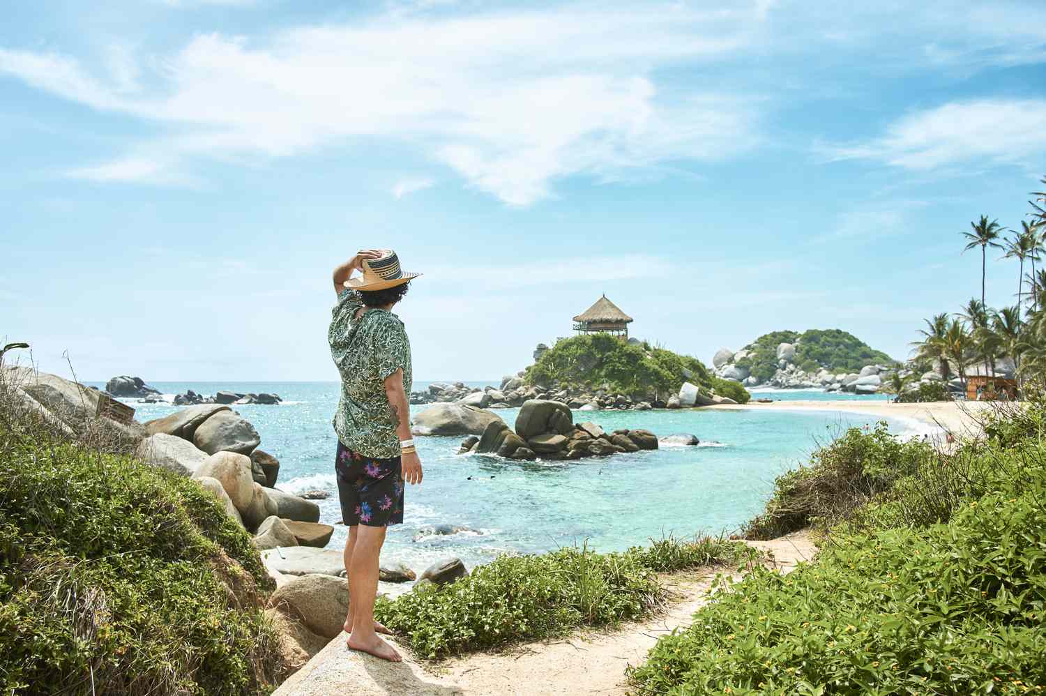 Person standing on a rocky beach looking towards a gazebo-like structure on a small island near a tropical coastline