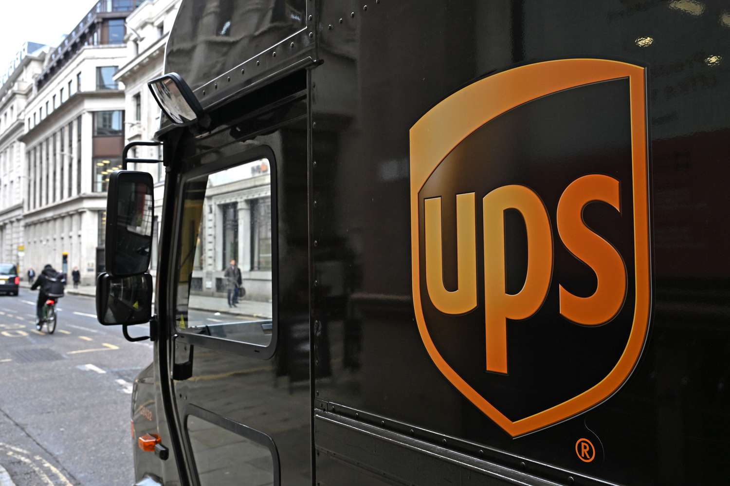 The UPS logo is seen on the side of a brown van, with people and buildings in the background.