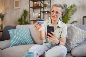 A woman sits on a couch, holding a bank card and mobile phone.