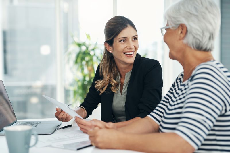 Young professional woman and an older woman sit in an office environment with a document and laptop.