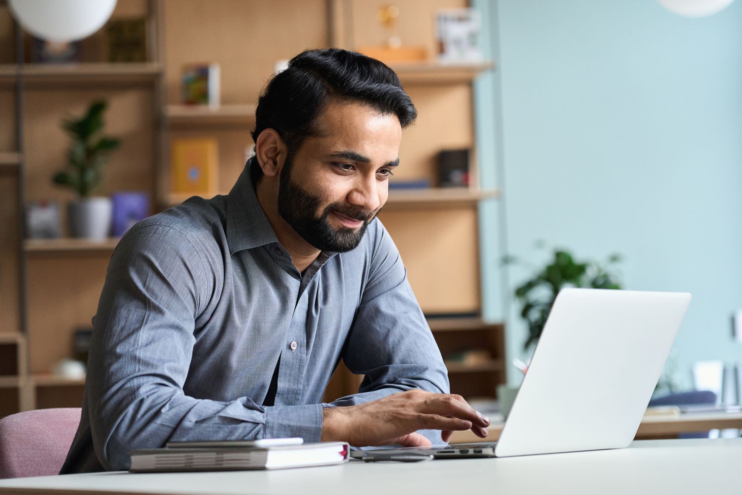A man in a grey button down shirt sits at a desk with a laptop