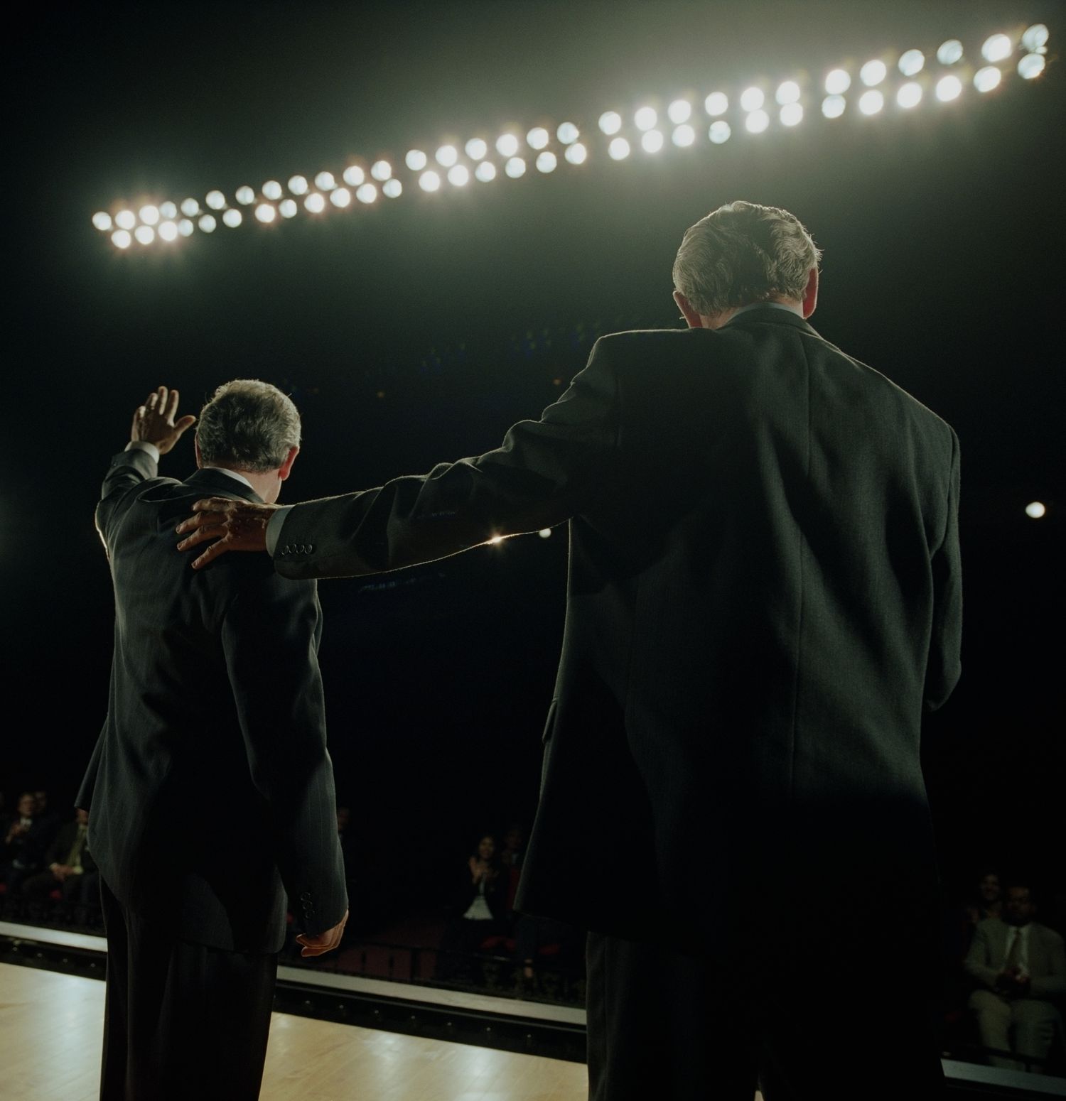 Two people in suits, with one waving to a ground from a stage in a darkened auditorium.