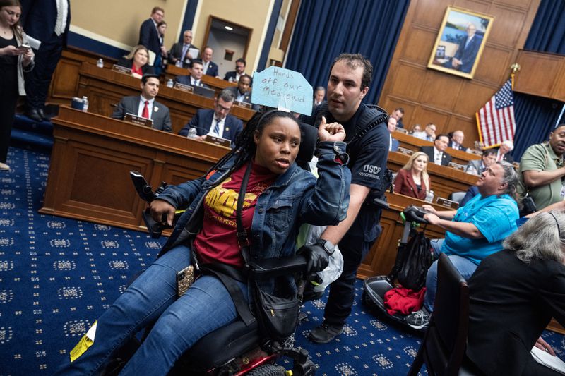 A protestor who has a sign attached to the back of their wheelchair is pushed by security from a meeting room as House Representatives watch. 