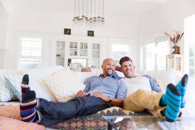 Comfortable male gay couple in striped socks relaxing, watching TV on living room sofa