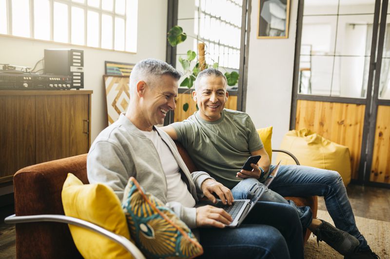 A male couple on the couch, one with a cell phone, the other with a laptop, going over their dividend returns.