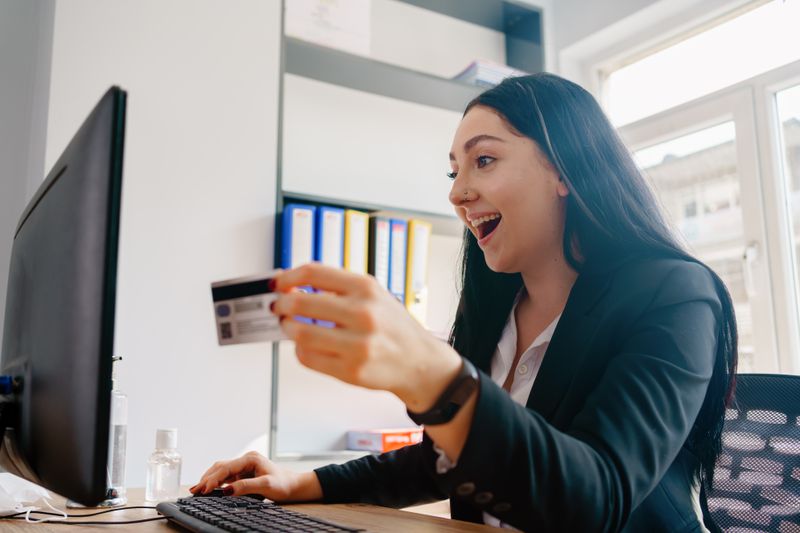 A happily surprised person holds a payment card while looking at her computer.