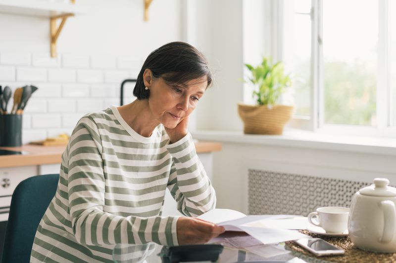 An older woman sits at a kitchen table looking at bills