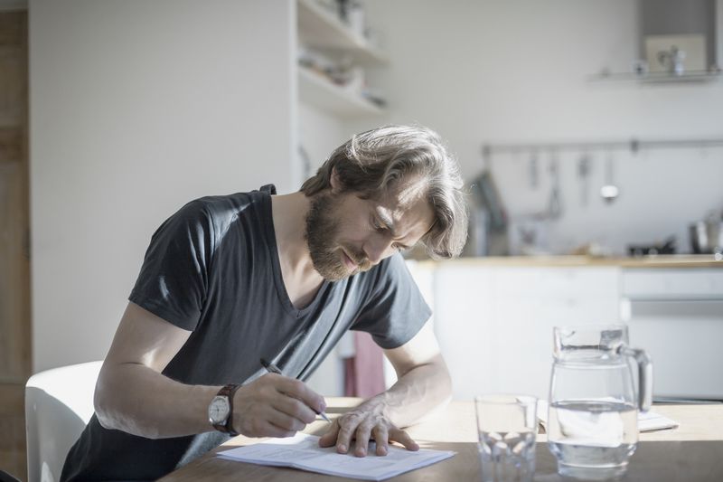 Man in casual clothing sitting at home writing on papers on the kitchen table