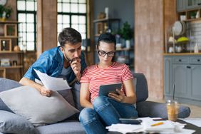 A couple sit together at a couch with a tablet computer and paperwork, analyzing their finances.