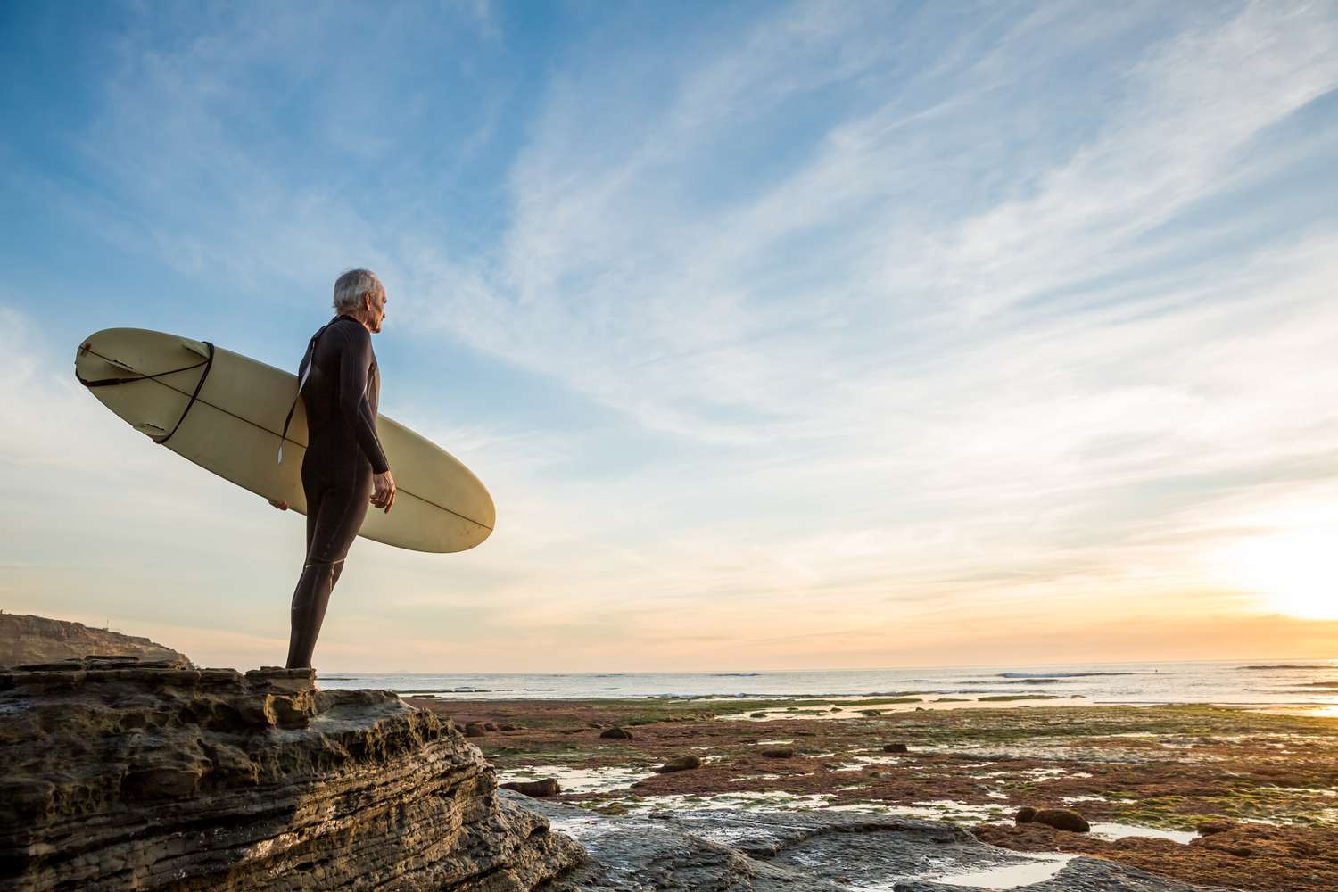 Senior retired surfer man looking at waves and holding a surfboard at Sunset Cliffs, San Diego, California.
