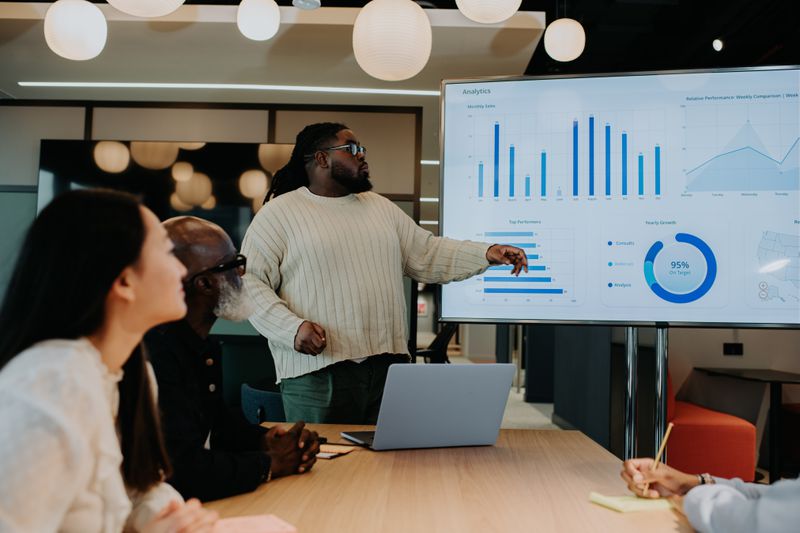 A meeting occurs in an office building. A man gives a presentation using a large monitor / screen. The screen displays graphs and charts. Colleagues listen intently.