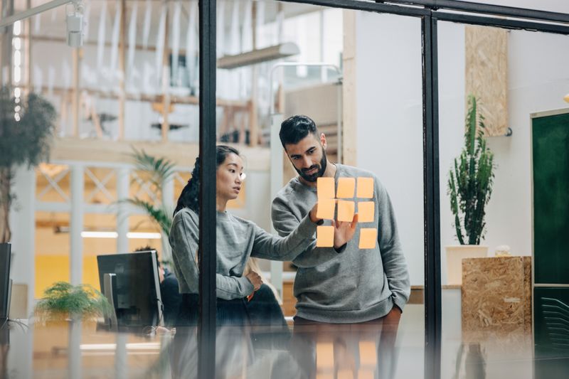 a man and woman talking while sticking notes on a glass wall in a modern office