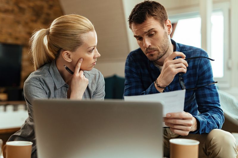Young couple sitting in their livign room and looking seriously at mortgage documents and a laptop