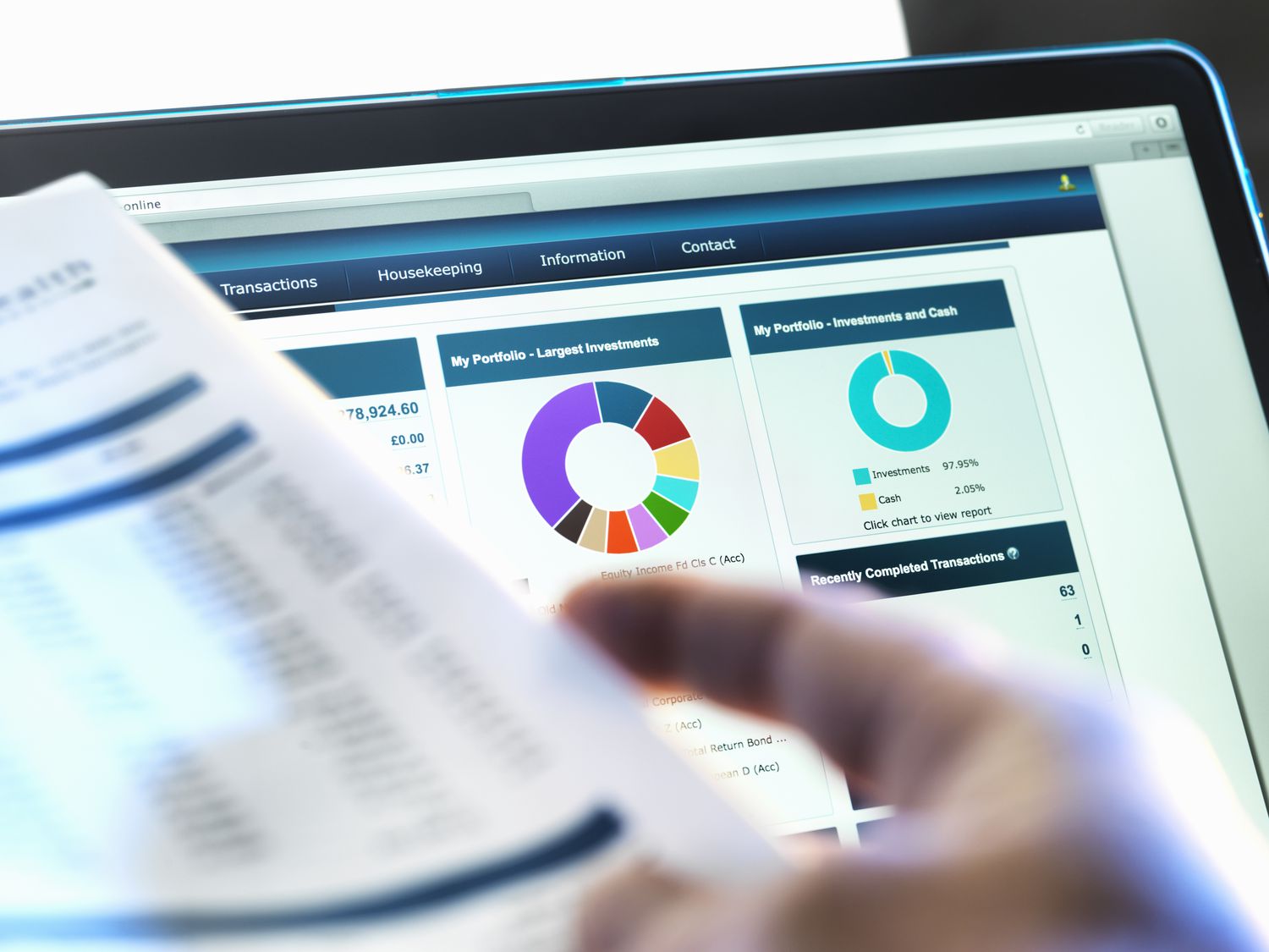 A computer screen close up showing colorful charts for an investment porfolio, while an out of focus hand in the foreground holds paperwork