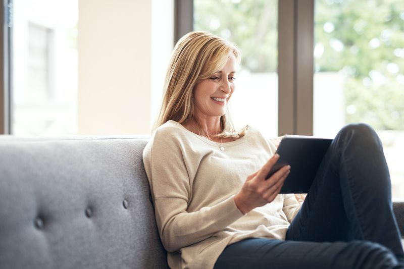 Woman in her 40s sitting on her sofa and looking happily at bank rates on her tablet