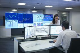 Financial analysts in an office with multiple computer monitors displaying charts and other data.