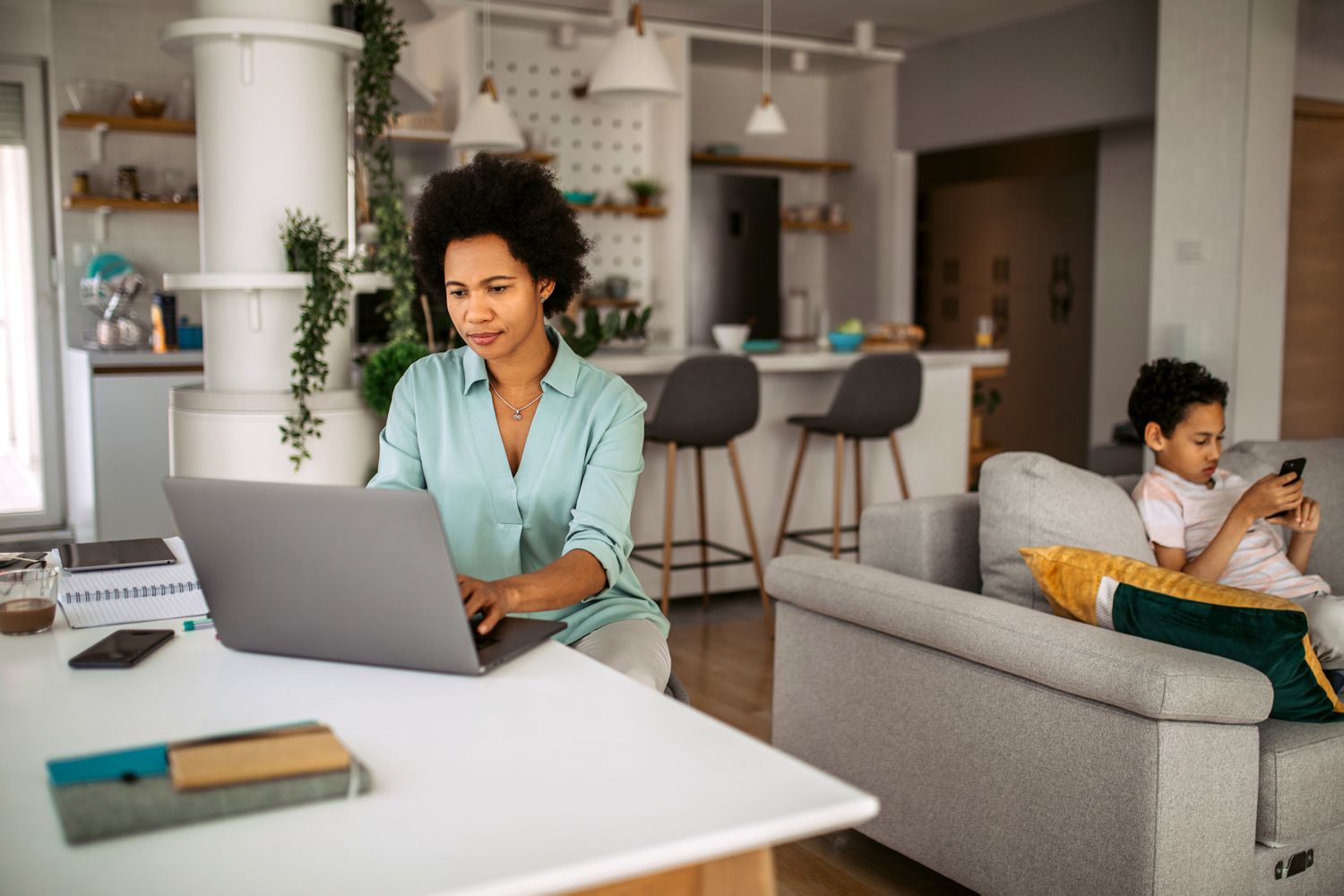 Mom in her 30s at her kitchen table, looking at a laptop while her child is on the couch