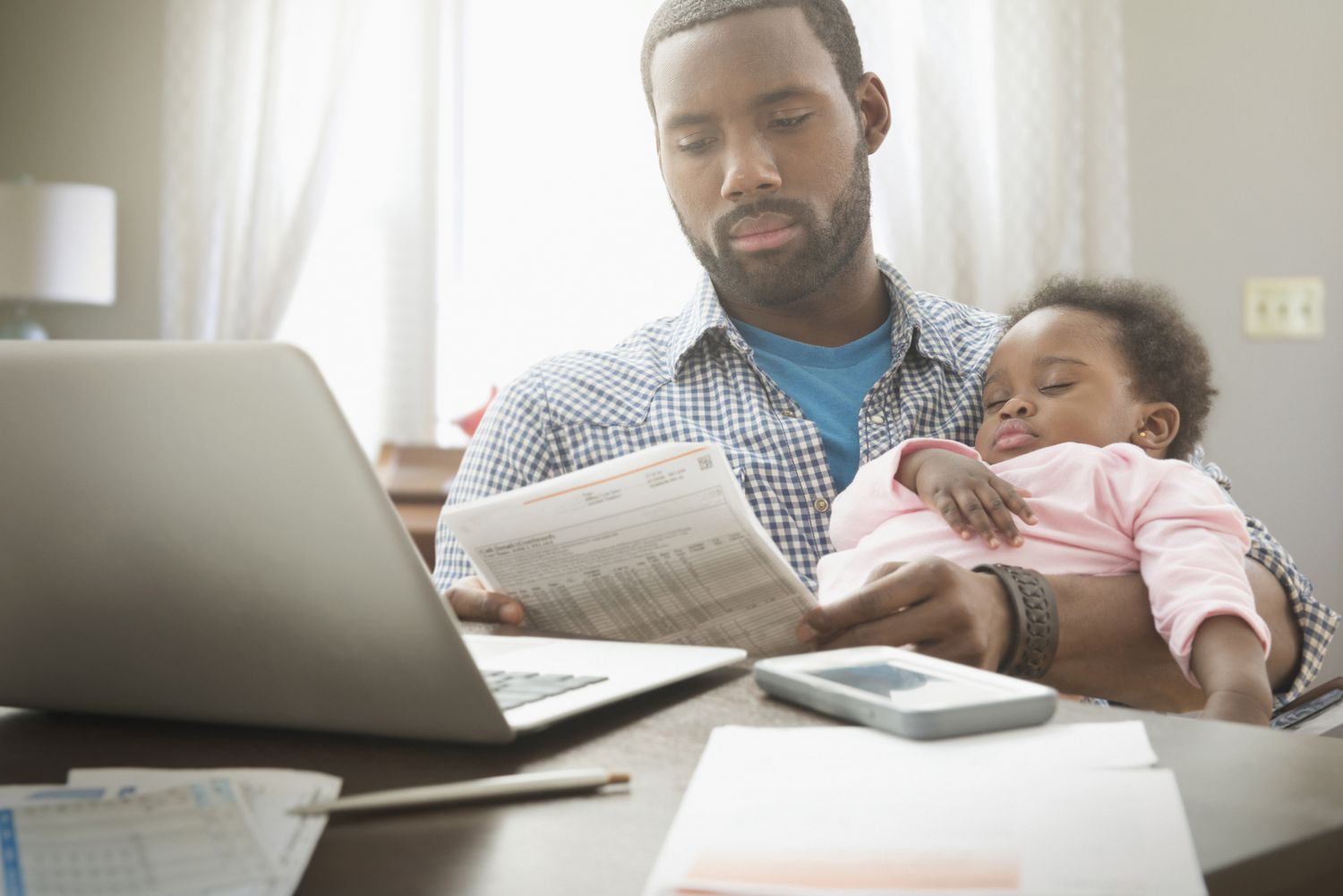 A father holds his sleeping baby while he looks at bills. 