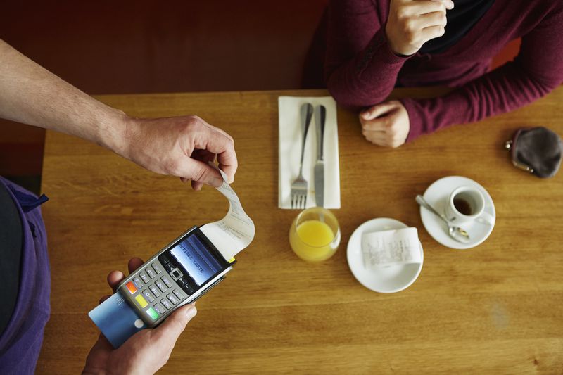 A waiter prints a credit card receipt using a handheld card reader for a customer seated at a cafe table.