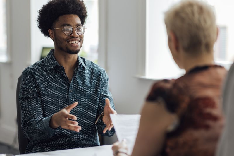 Over the shoulder view of a young male employee engaging in a focused discussion with a mature female manager at the managers desk in an office.